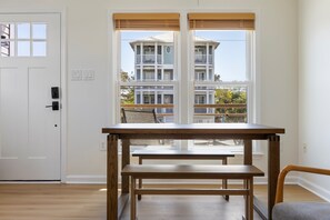 Una habitación con una mesa de madera y un banco, una puerta blanca y ventanas amplias con vistas a un edificio de varios pisos.