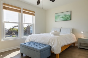 Un dormitorio con cama, ventana con persianas, mesita de noche con lámpara y un cuadro en la pared.