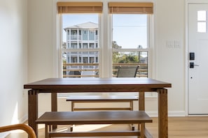 Una habitación con una mesa y banco de madera, una puerta blanca y vista a un edificio a través de una ventana con persianas.