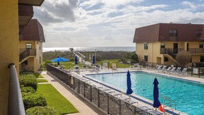 Uma piscina com espreguiçadeiras e guarda-sóis, vista para o oceano e um edifício com varanda.