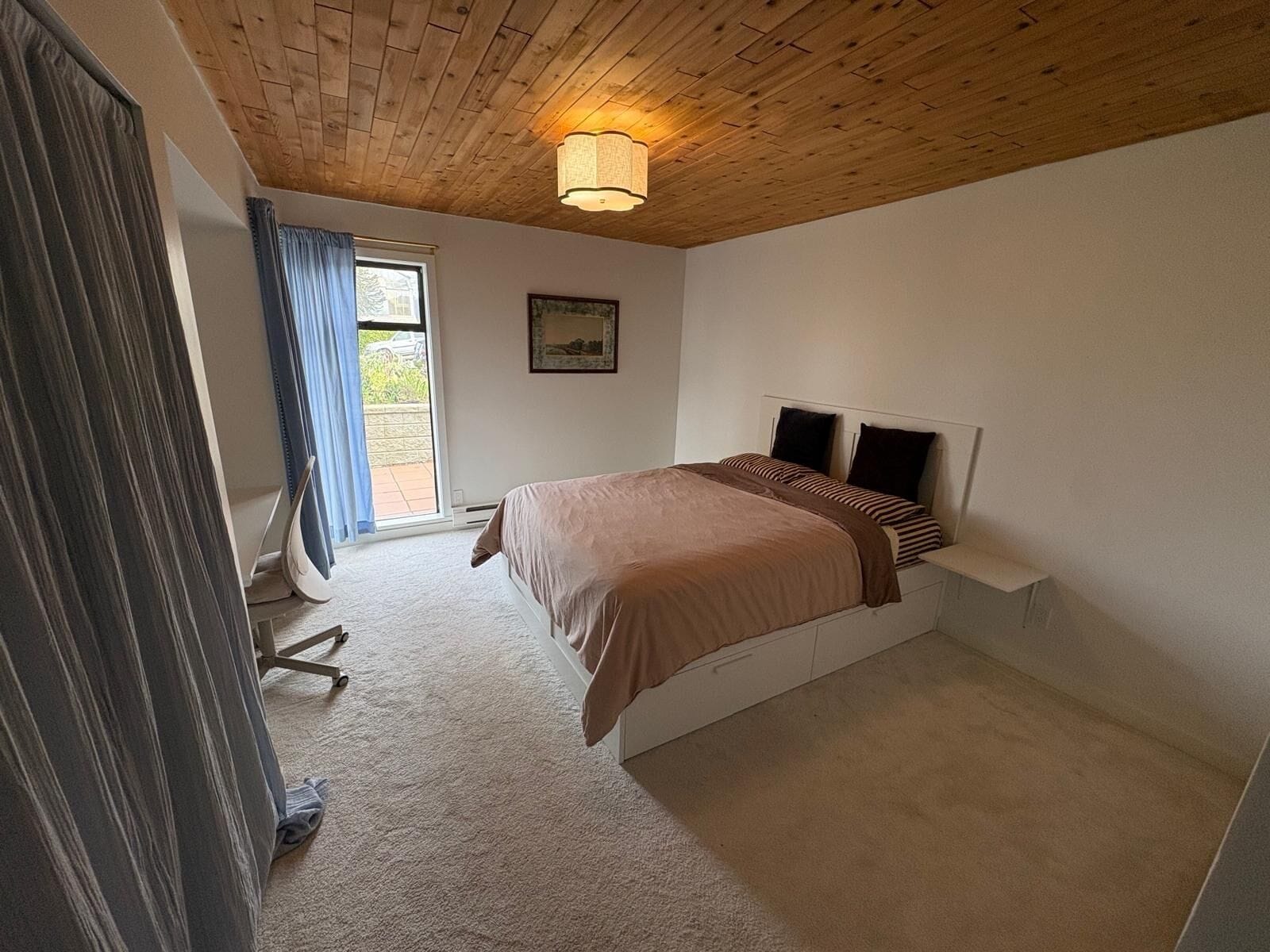 Ein Schlafzimmer mit Holzboden, einem Bett mit Kopfteil, einem Fenster mit Vorhängen und einem kleinen Nachttisch.