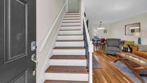 A staircase with white steps and a dark door, leading to a living area with a sofa, a coffee table, and a dining table. - Great Home_Comfy Beds_Sleeps 8_Hot Tub & Game Room (Kansas City)