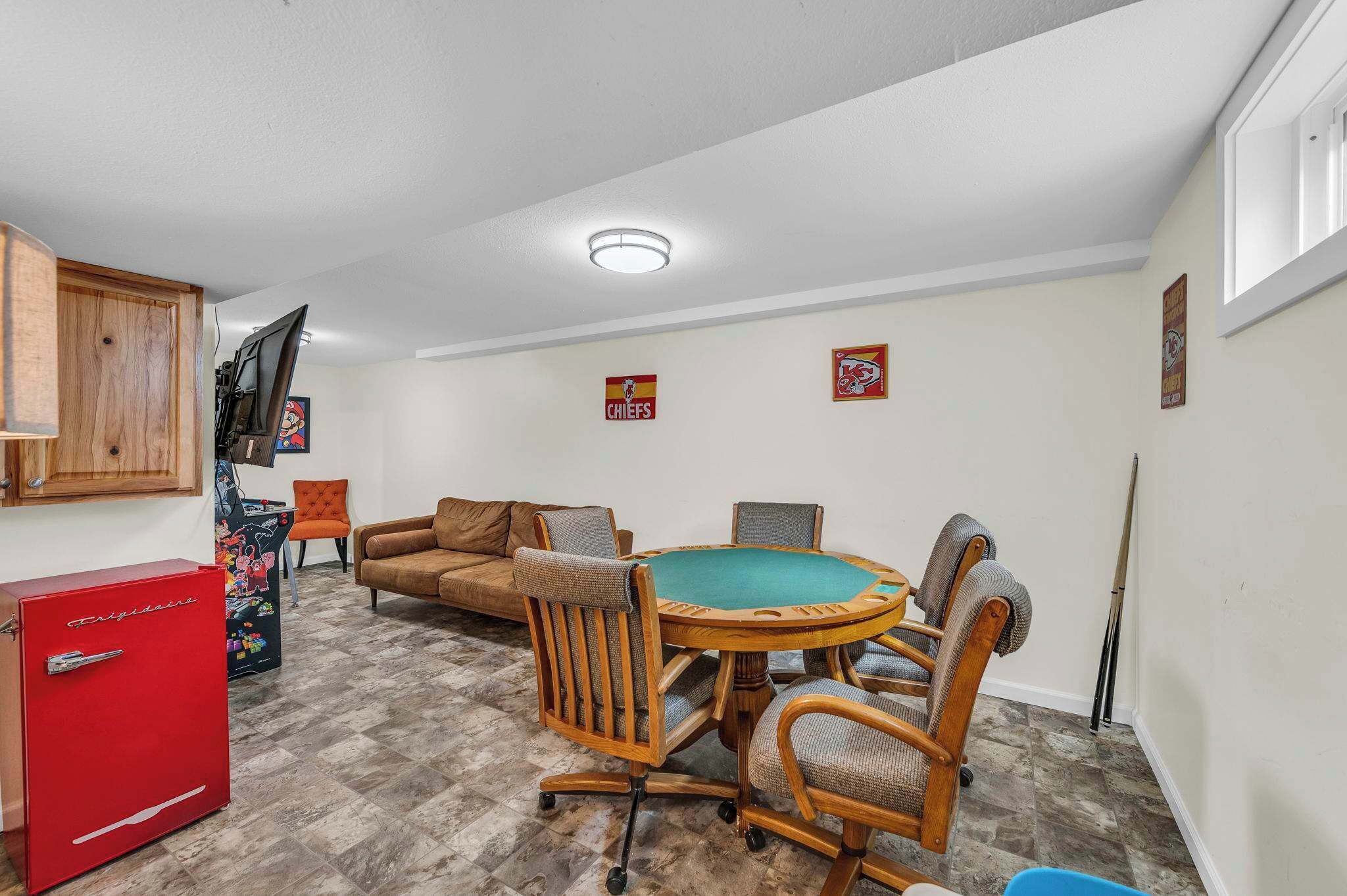 A room with a pool table, a red refrigerator, a wooden cabinet, a couch, and two framed sports posters on the wall.