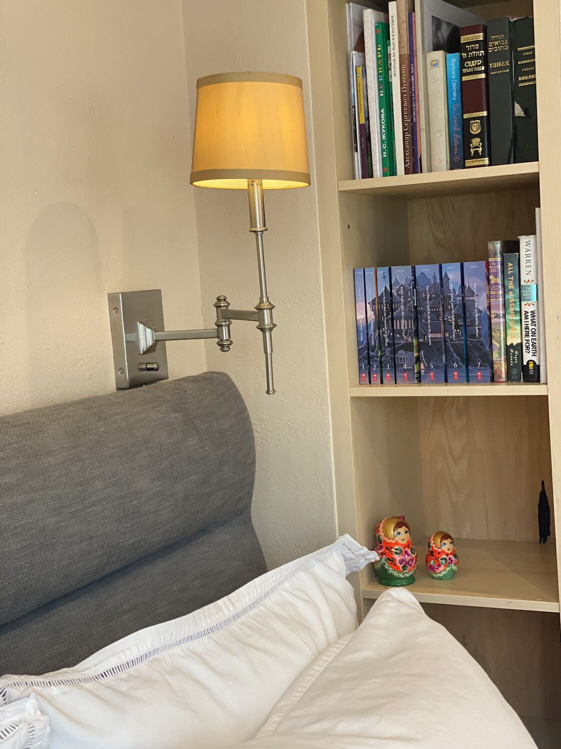 Ein Bett mit weißer Decke, ein graues Sofa, eine Wandlampe und ein Bücherregal mit Büchern und Dekoration.