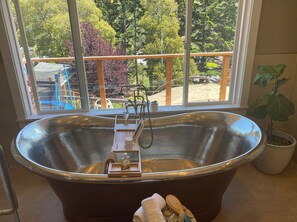 Une grande baignoire sur pieds, avec un plateau en bois sur lequel sont posés une bouteille et une tasse, une serviette et une brosse.
