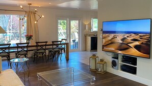 Uma sala de estar moderna com uma grande televisão de tela plana, piso de madeira, uma mesa de centro de vidro e um espaço de jantar com cadeiras e uma mesa.
