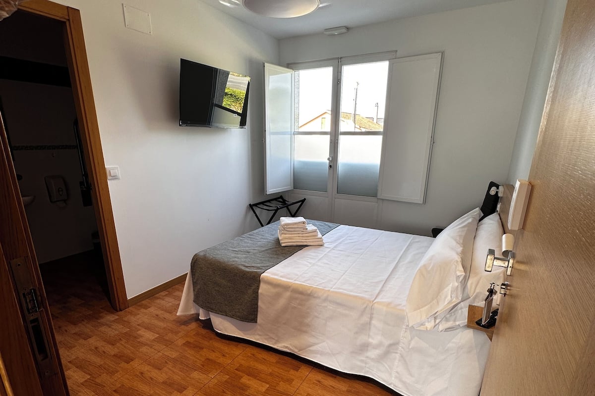 Un dormitorio con cama, ventilador de techo, televisor montado en la pared y ventana con vistas.