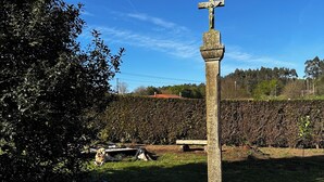 A cross monument in a grassy area with a hedge and trees in the background. - Pensión Mirador de Rourís (O Pino)