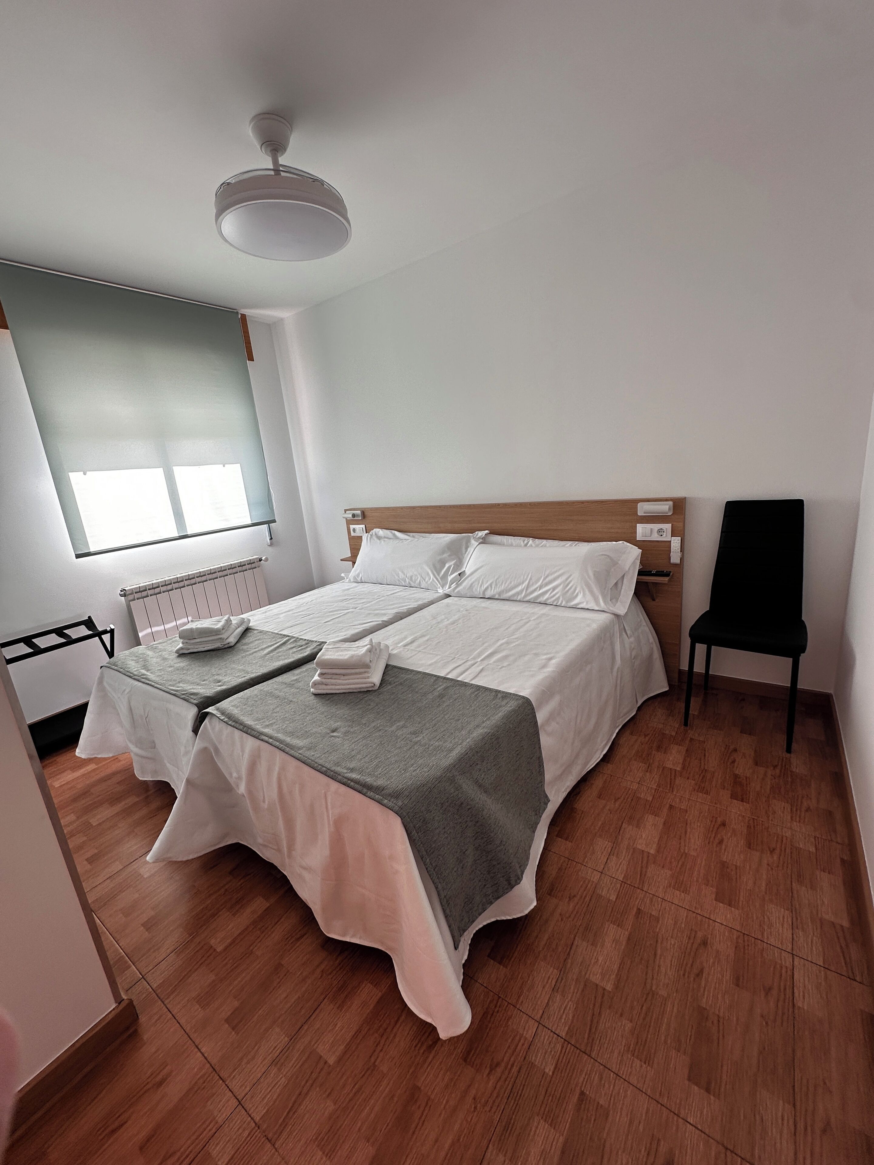 A neatly made bed with white linens and a grey blanket, a black chair, and a ceiling fan in a room with wooden flooring and a window with blinds.