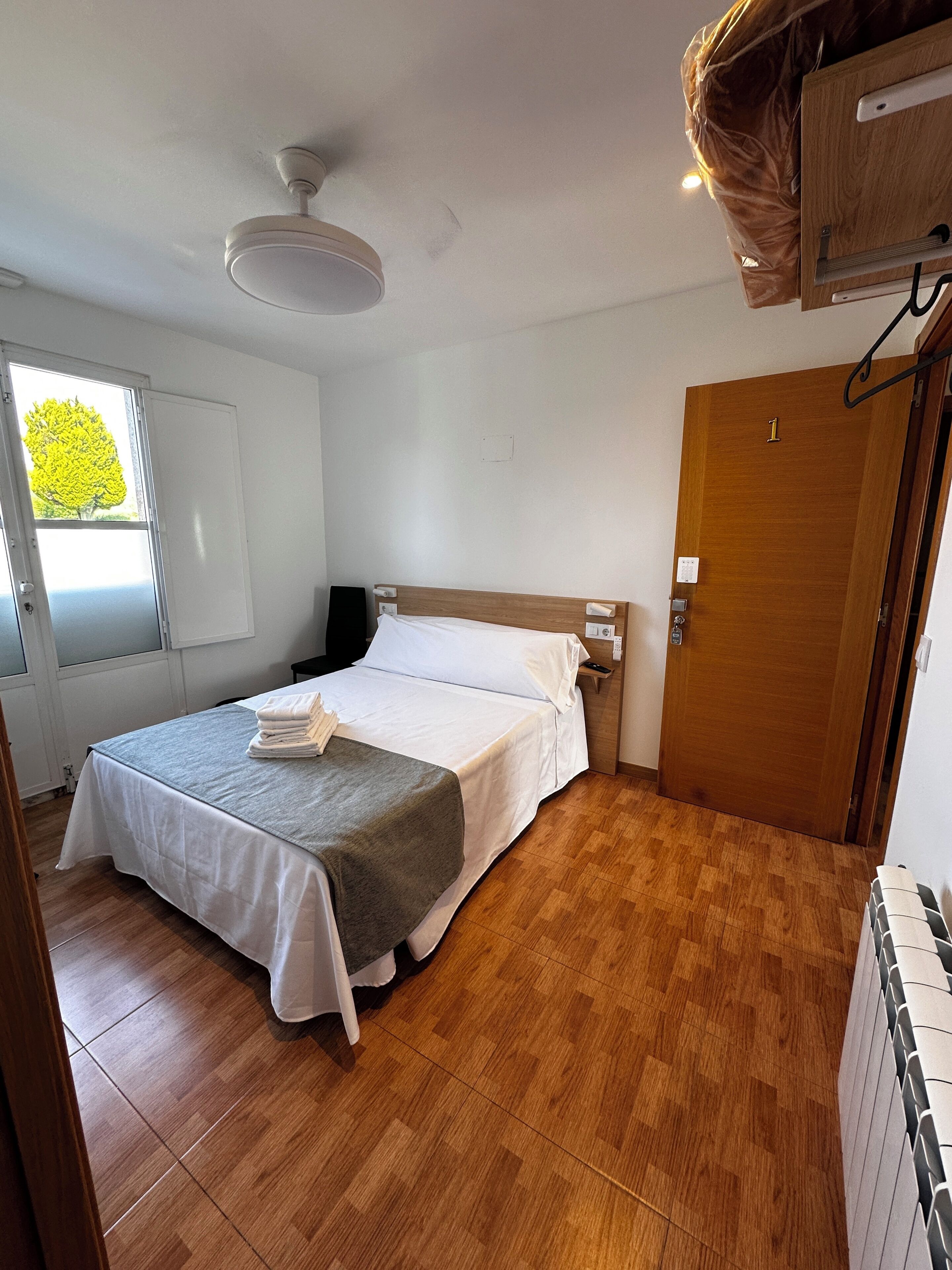 A single bed with a grey bedspread, a wooden headboard, a bedside table with a lamp, a window with a view of trees, and a wooden door.