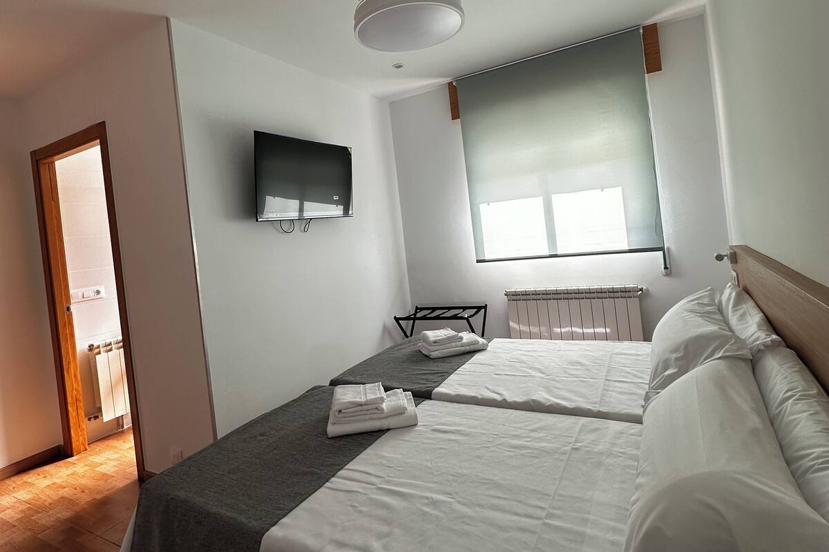Habitación de hotel con cama, televisor montado en la pared, ventana con persianas y ventilador de techo.