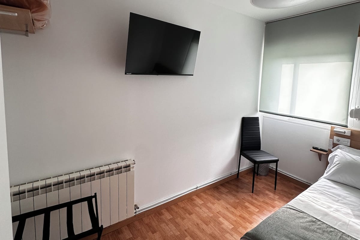 Un dormitorio con cama, una silla, televisor montado en la pared, radiador y ventana con vistas.