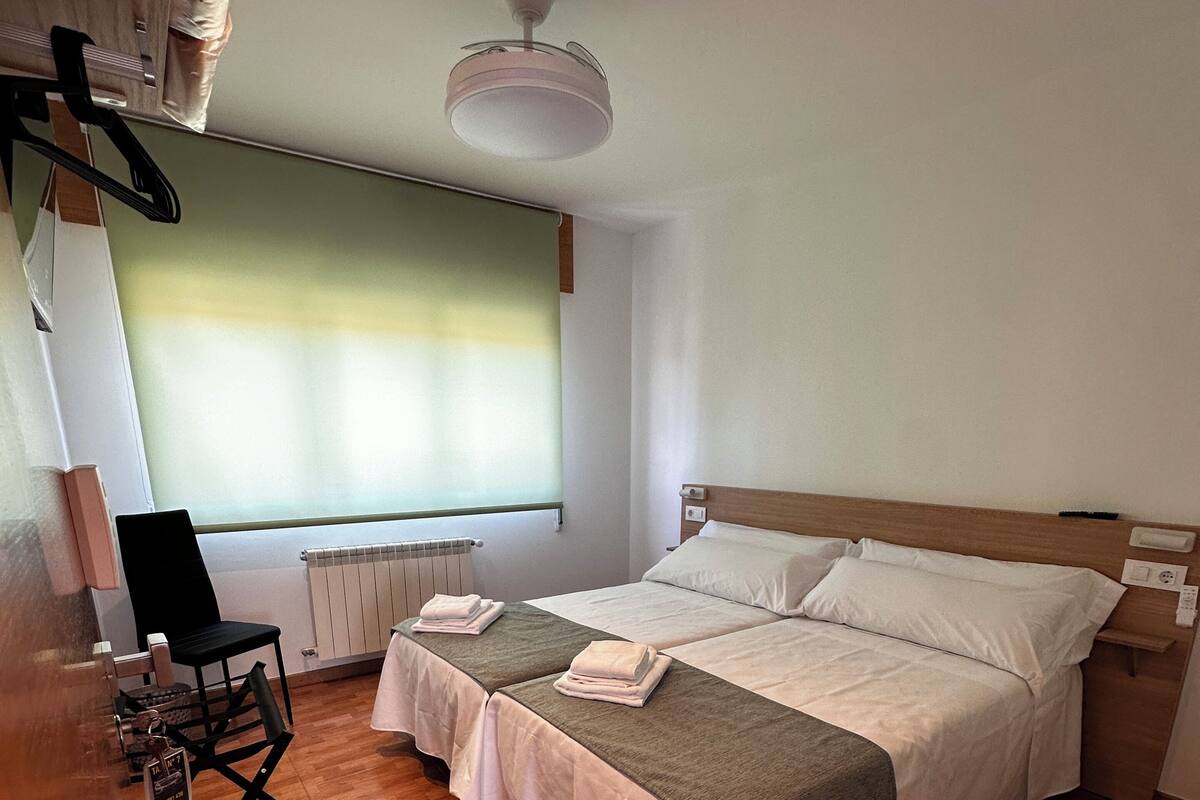 Habitación de hotel con suelo de madera, una cama con cabecera, una mesita de noche, una silla, una ventana con persianas y un ventilador de techo.