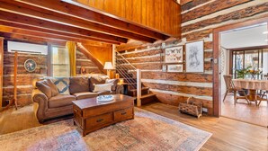 Interior de una cabaña de troncos con escalera de madera, sofá, mesa de centro y zona de comedor.