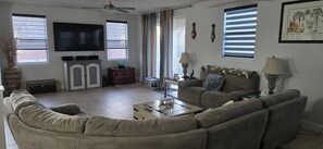 A living room with a sofa, a television, a ceiling fan, and a window with blinds.