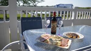 Una mesa de vidrio con una botella de vino, dos copas y un plato de comida en un balcón con valla blanca y vista a una casa y árboles.