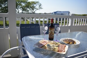 Ein Glastisch mit einer Weinflasche, zwei Weingläsern und einem Teller Essen auf einem Balkon mit weißem Geländer und Blick auf ein Haus und Bäume.