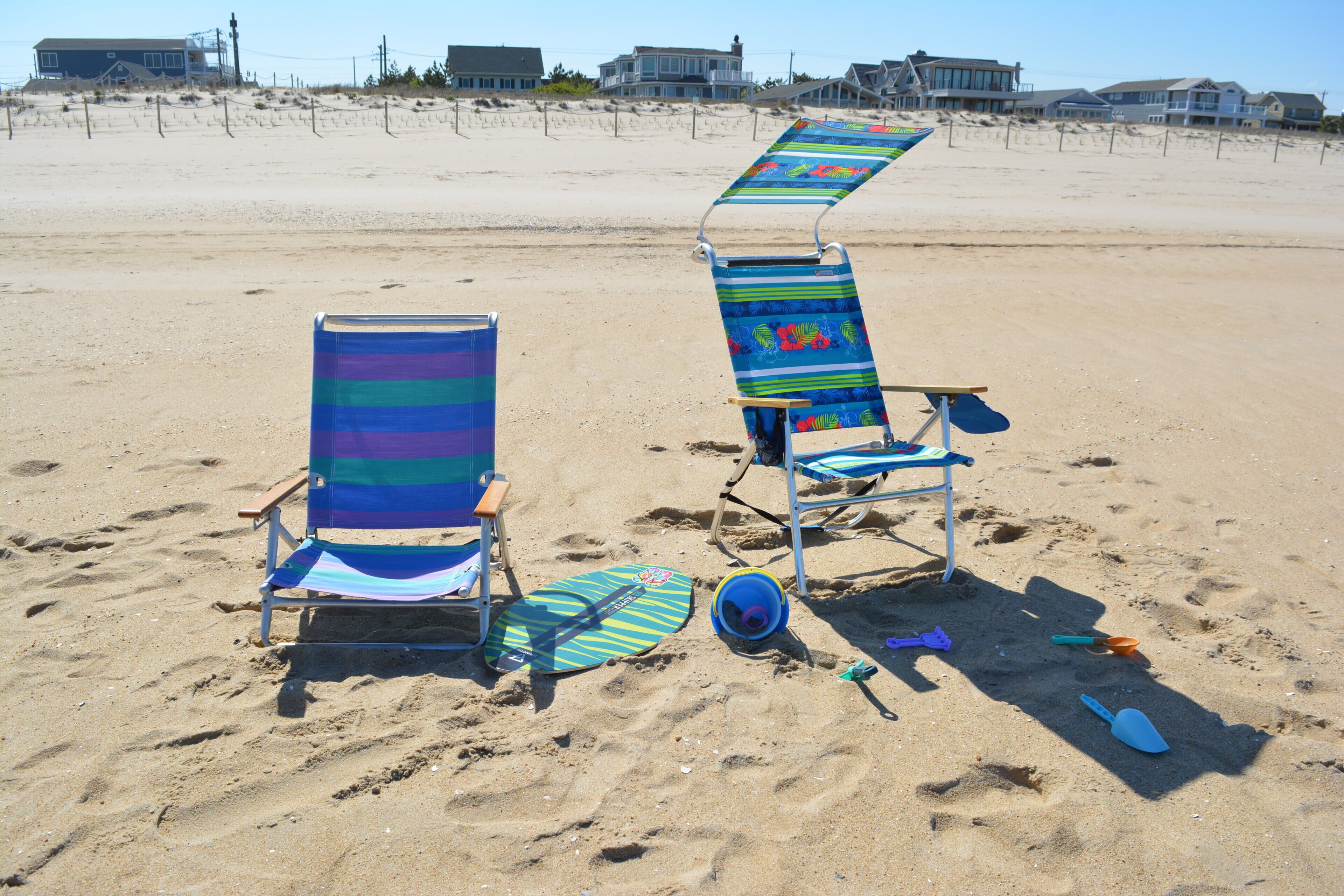 Two beach chairs with a beach umbrella, a pair of flip-flops, and a blue shovel on a sandy beach with houses in the background.