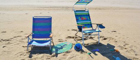 Two beach chairs with a beach umbrella, a pair of flip-flops, and a blue shovel on a sandy beach with houses in the background.
