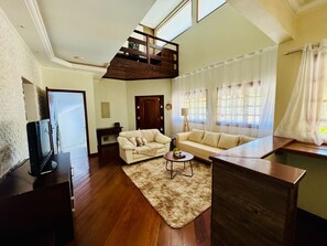 A spacious living room with a high ceiling, wooden flooring, a sofa set, a coffee table, and a television.