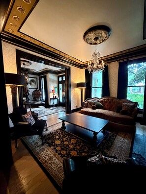A spacious living room with a large sofa, a coffee table, and a chandelier. There is a view of the outdoors through large windows. - "THE MAYBELLE" 1892 Historic Two Story Victorian Home, Downtown Springfield, TN (Springfield)