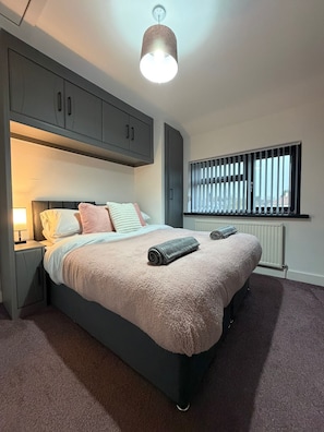 A bedroom with a bed, a nightstand, a window with blinds, and a ceiling light fixture.