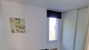 A small, single-bed room with a green bedspread, a window with blinds, a radiator, and a wall-mounted picture. - 247 - Your stay facing Hourtin Port – Superb view and peaceful setting (HOURTIN)