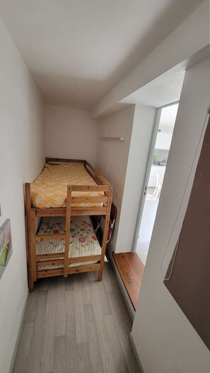 Ein kleines Zimmer mit einem Einzelbett, einem Holtzrahmen, einem Fenster und einer Tür, die auf einen Balkon führt.