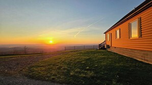 Une cabane en bois, avec des fondations en briques, une clôture en bois et une vue sur le coucher de soleil au-dessus d’un paysage vallonné.