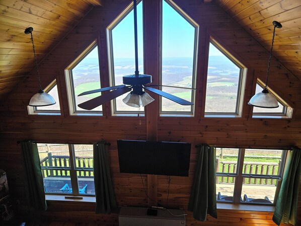 A room with a sloped ceiling, large windows, a ceiling fan, and a flat-screen TV.