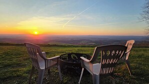 Deux chaises et un foyer sur une colline herbeuse, avec une vue sur le coucher de soleil.