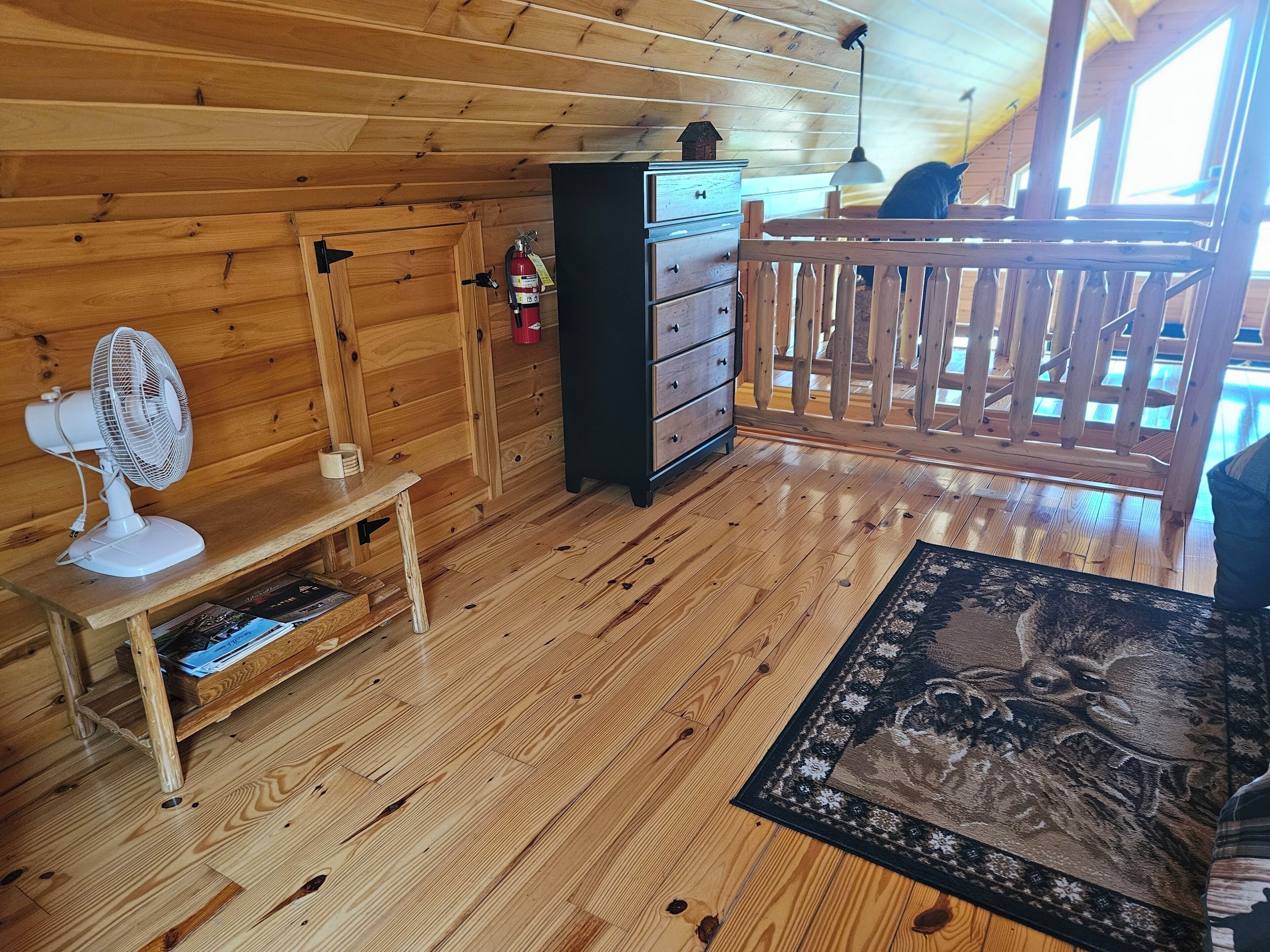 Interior de una cabaña de madera con un ventilador, cómoda y vistas a través de una baranda de madera.