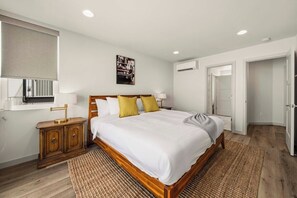 A bedroom with a wooden bed, white bedding, a bedside table with lamps, a wall-mounted air conditioner, and a door leading to another room. - Sleek 3BR Seattle Townhome - Near UW-Husky Stadium (Seattle)