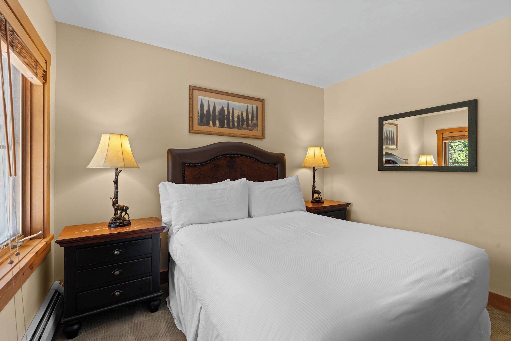 A neatly made bed with white linens, two bedside lamps, a wooden nightstand, a framed picture on the wall, and a mirror above the bed.