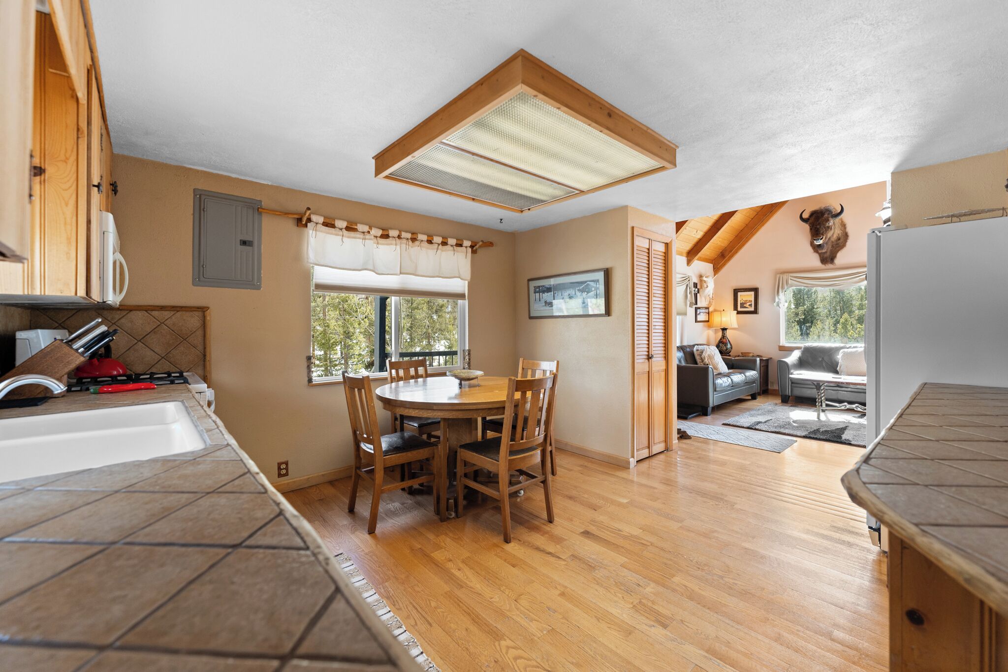 A kitchen with a dining table, chairs, and a ceiling fan. There is a window with a view of trees and a mounted animal head on the wall.
