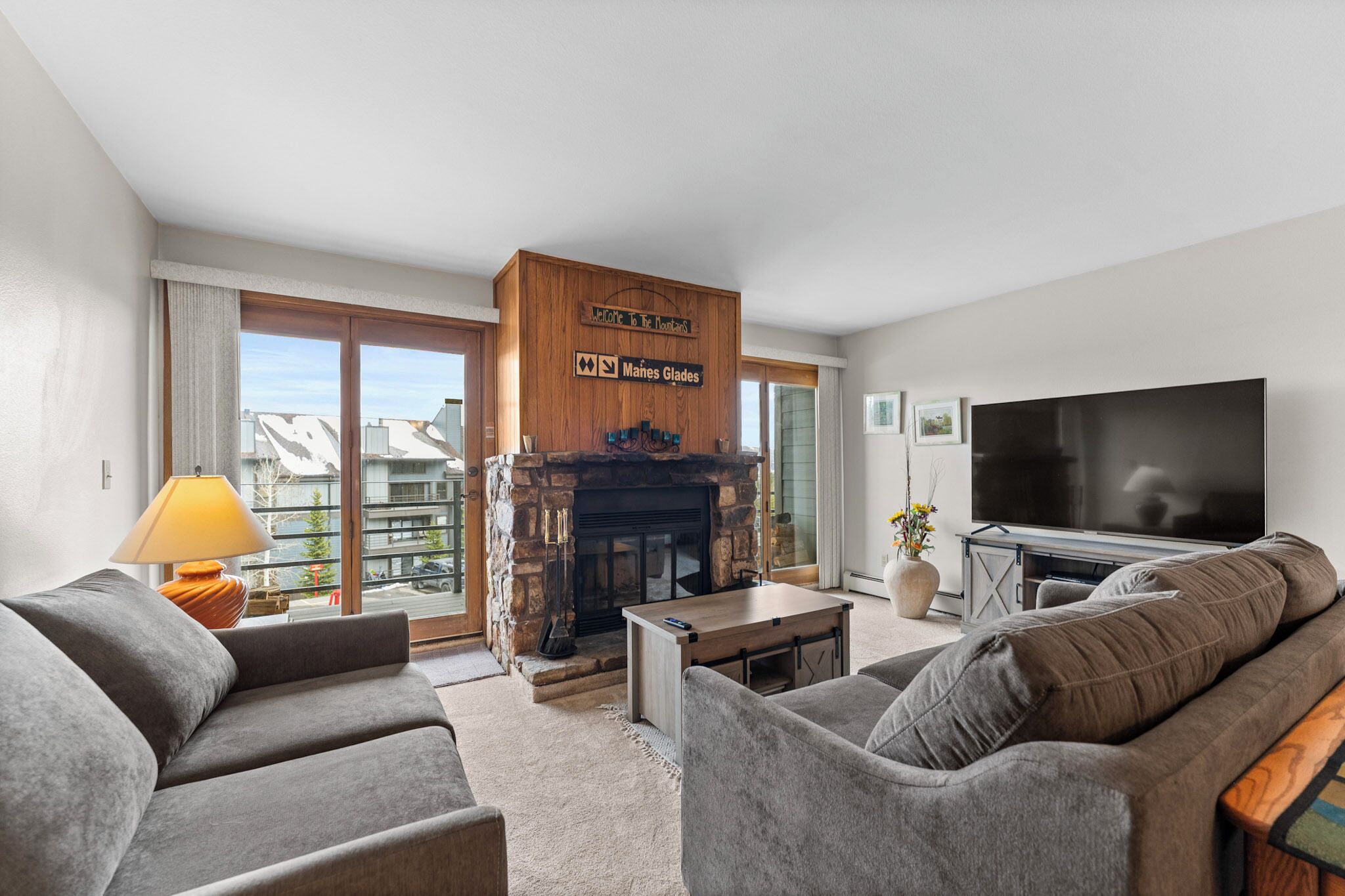 A modern living room with a stone fireplace, grey sofas, a flat-screen TV, and a balcony with a view.