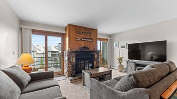 A modern living room with a stone fireplace, grey sofas, a flat-screen TV, and a balcony with a view.