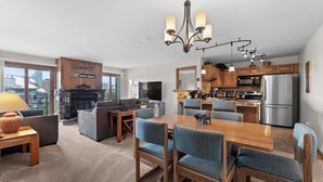 A modern living room with a fireplace, a dining area with blue chairs, and a kitchen with wooden cabinets and stainless steel appliances. - Brv203aa (Silverthorne)