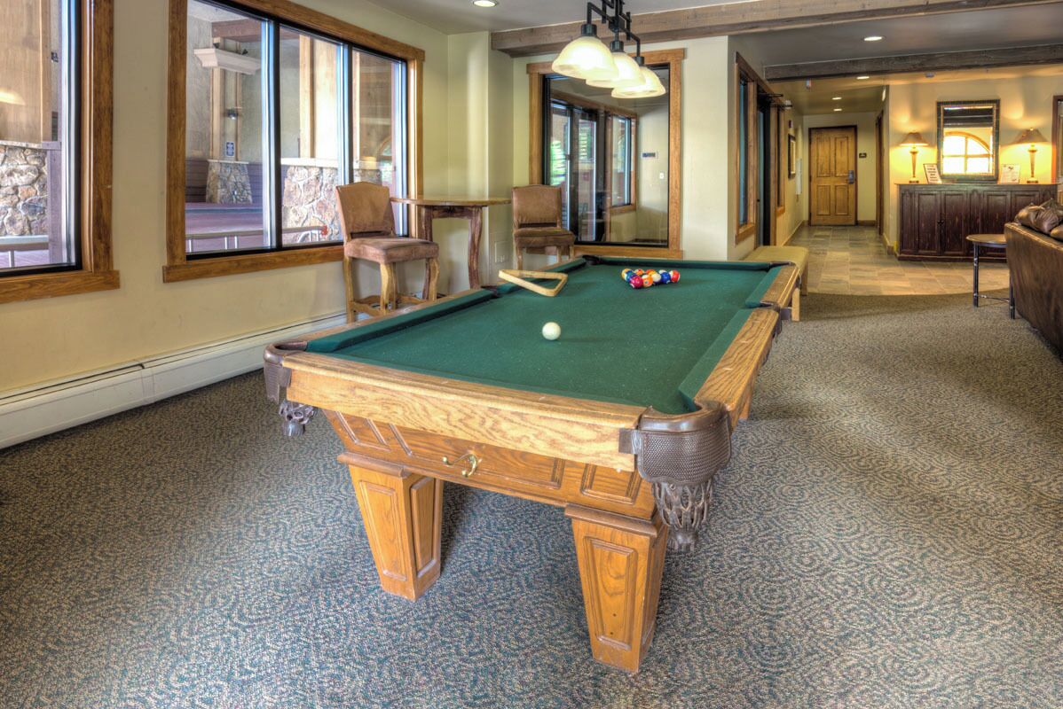 A pool table in a room with large windows, a wooden bench, and a small table with a lamp.