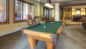 A pool table in a room with large windows, a wooden bench, and a small table with a lamp. - Red Hawk Lodge #2212 (Keystone)
