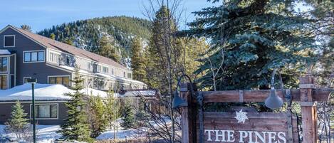 A wooden sign with "THE PINES" written on it, surrounded by snow-covered ground and trees.