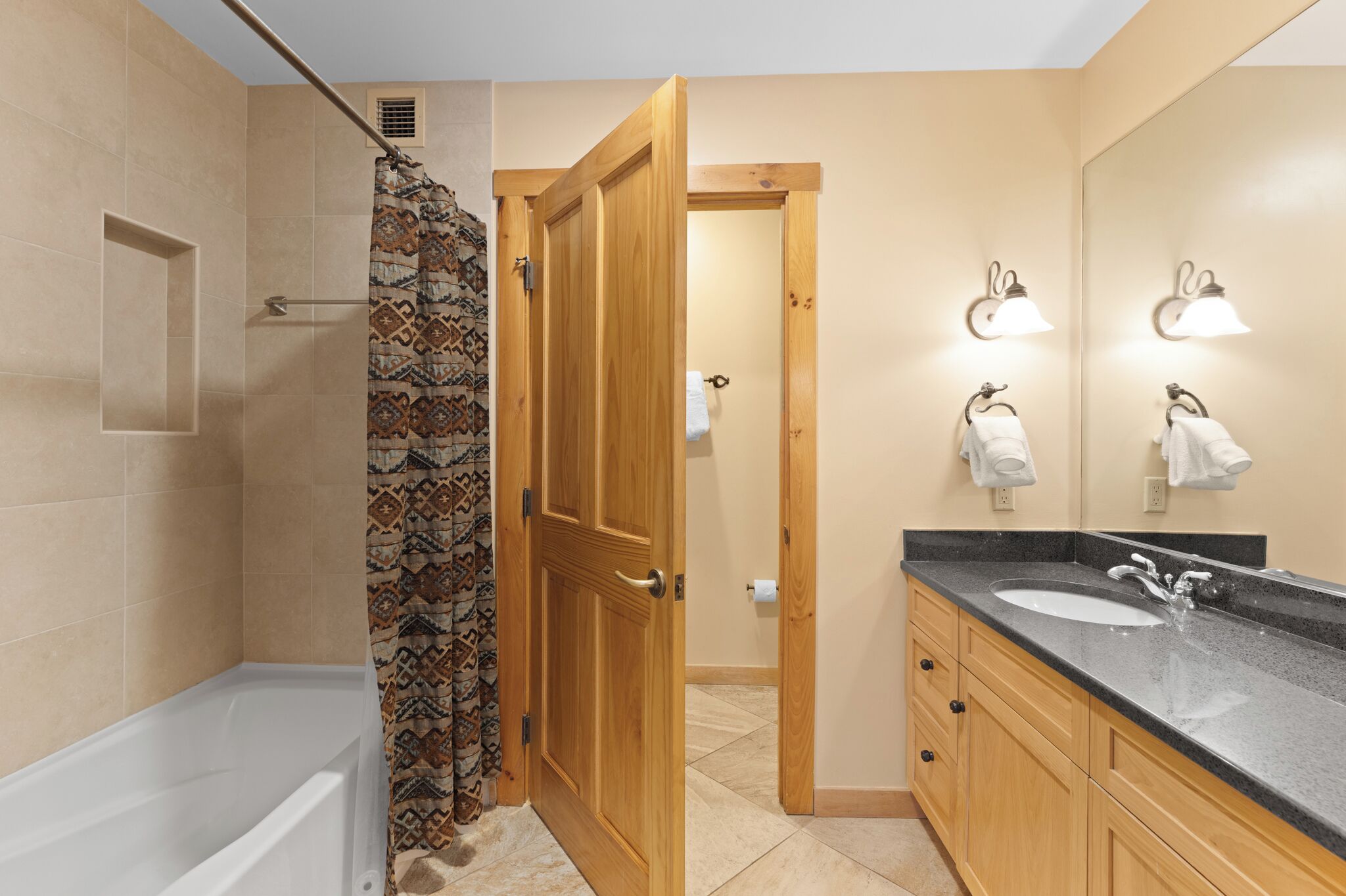 Un baño con bañera, cortina de ducha, puerta de madera, tocador con lavabo y espejo.