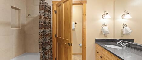 Un baño con bañera, cortina de ducha, puerta de madera, tocador con lavabo y espejo.