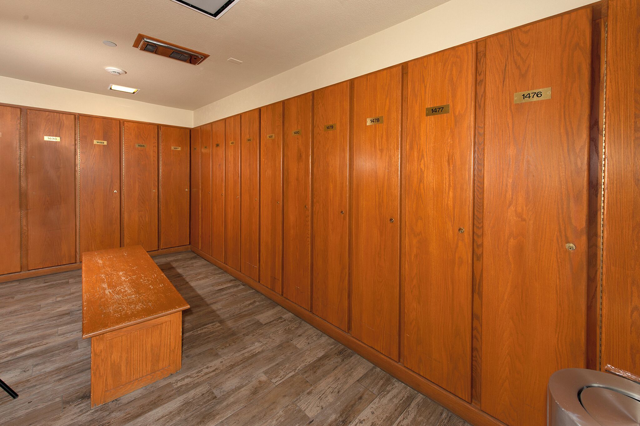 A row of wooden lockers with numbers on them, a bench, and a trash can.