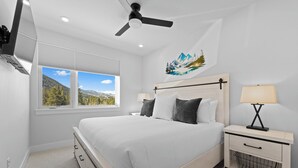 Un dormitorio con cama, mesita de noche, lámpara, ventilador de techo y ventana con vista a la montaña.