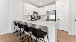 Una cocina moderna con muebles blancos, un refrigerador de acero inoxidable, un microondas y una cafetera. Hay tres taburetes negros con asientos de cuero.