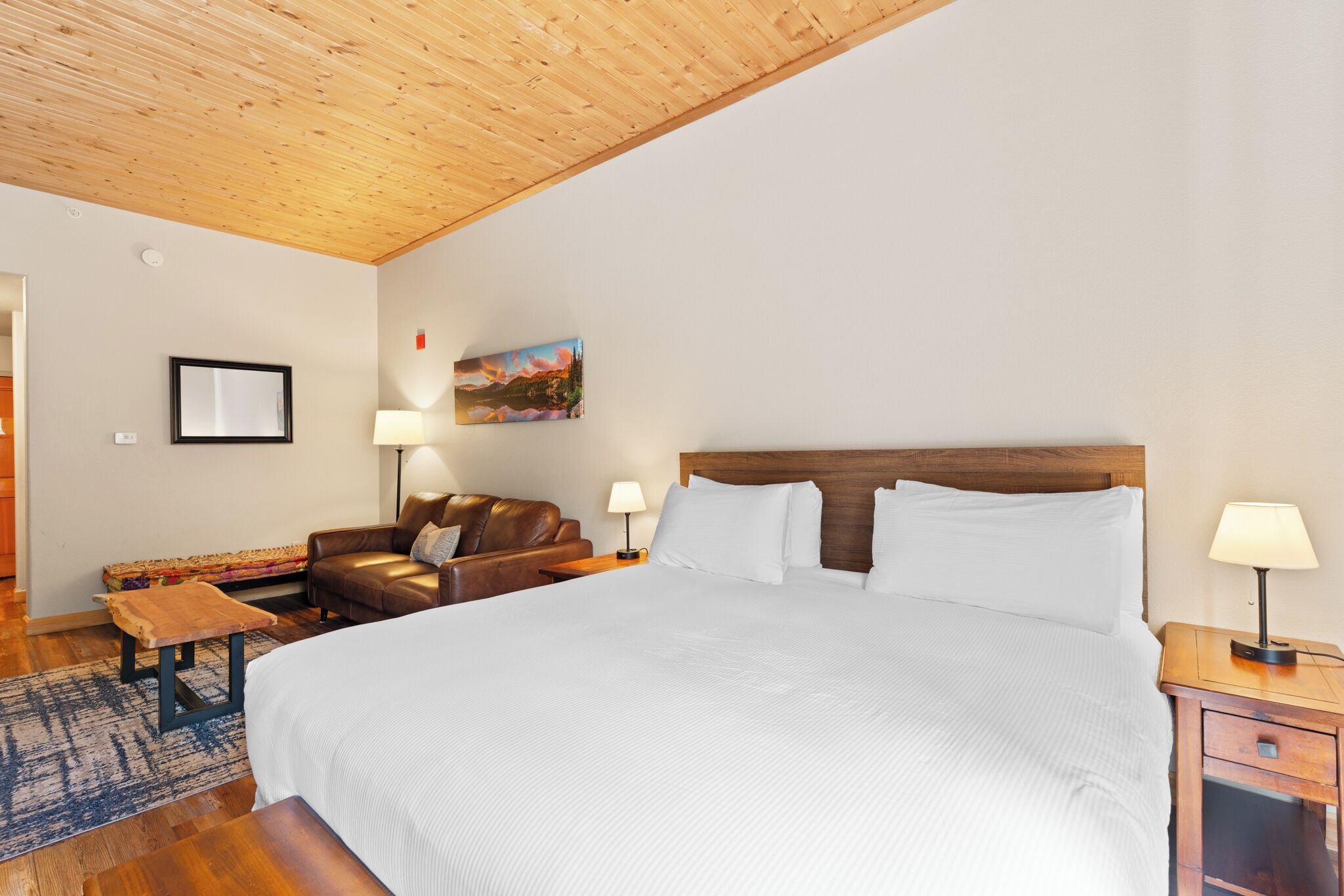 A hotel room with a wooden ceiling, a large bed with white bedding, a brown leather sofa, a small wooden table, and two bedside lamps.
