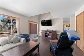 A living room with a sofa, a dining table, a TV mounted on the wall, and a view of the outdoors. - 204efc-2 (Silverthorne)