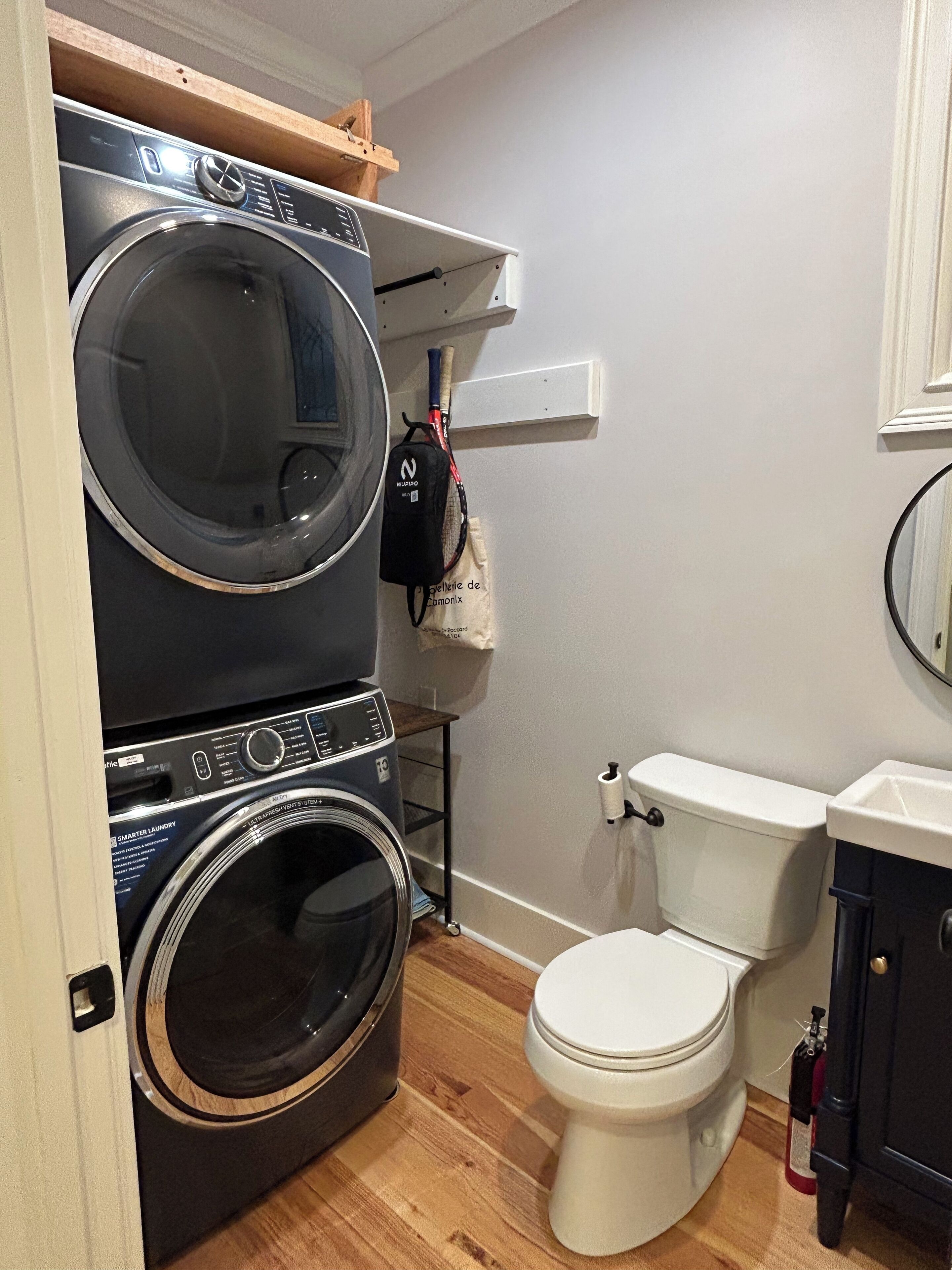 A compact laundry room with a stacked washer and dryer, a toilet, and a small shelf with a bag.