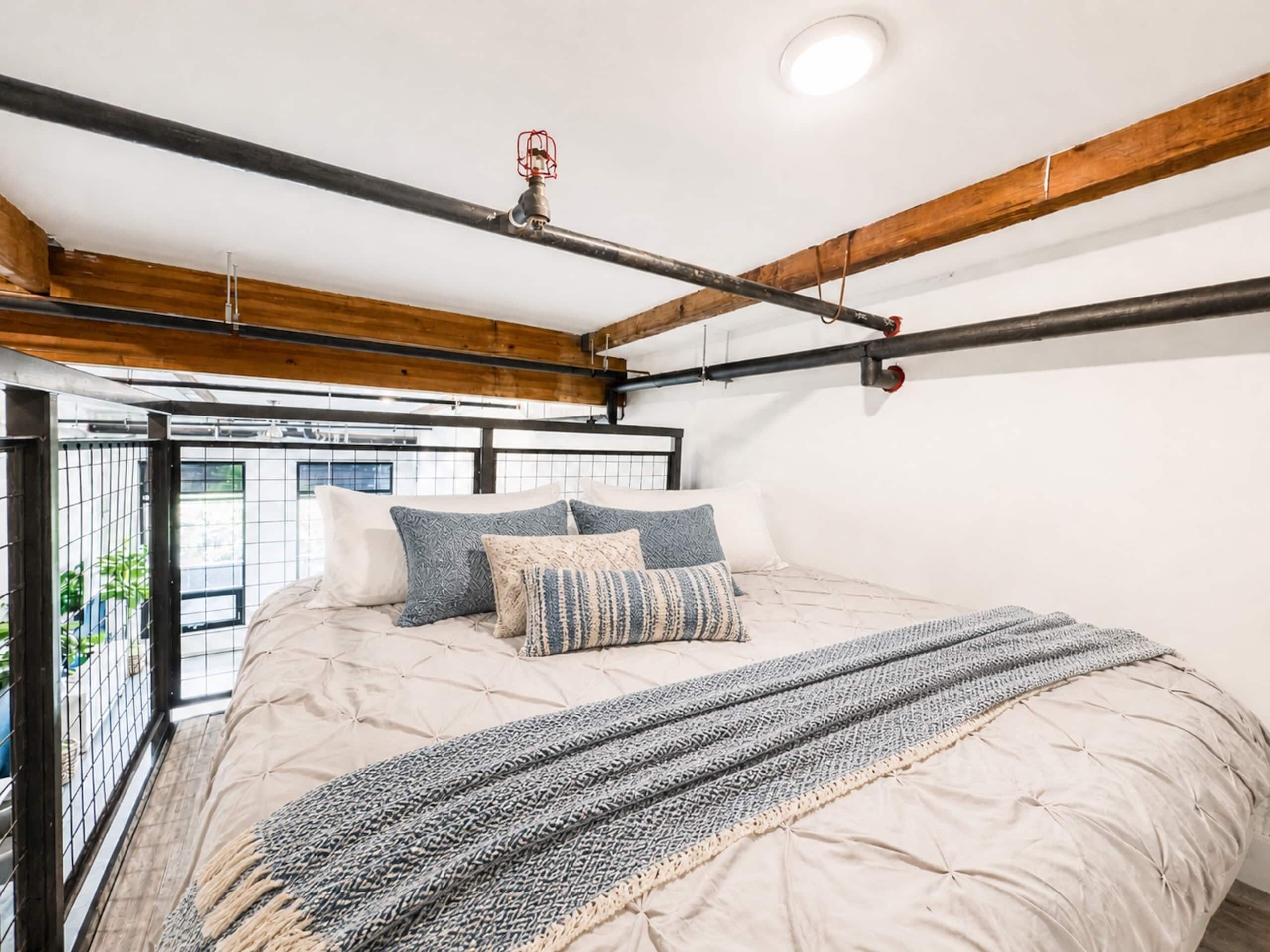 Un dormitorio con cama, almohadas, una colcha, vigas de madera a la vista y una ventana con vistas.